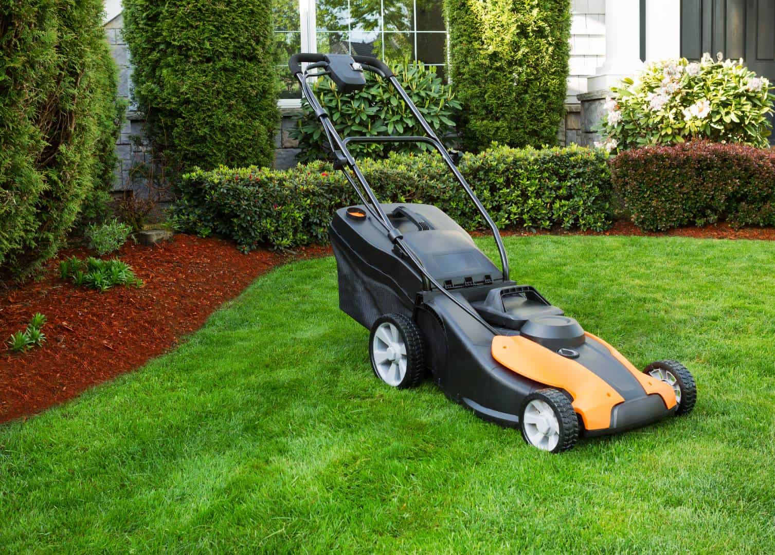 Orange and black electric lawn mower sits on a vibrant green lawn with trimmed hedges and mulch bed near a home's front windows.