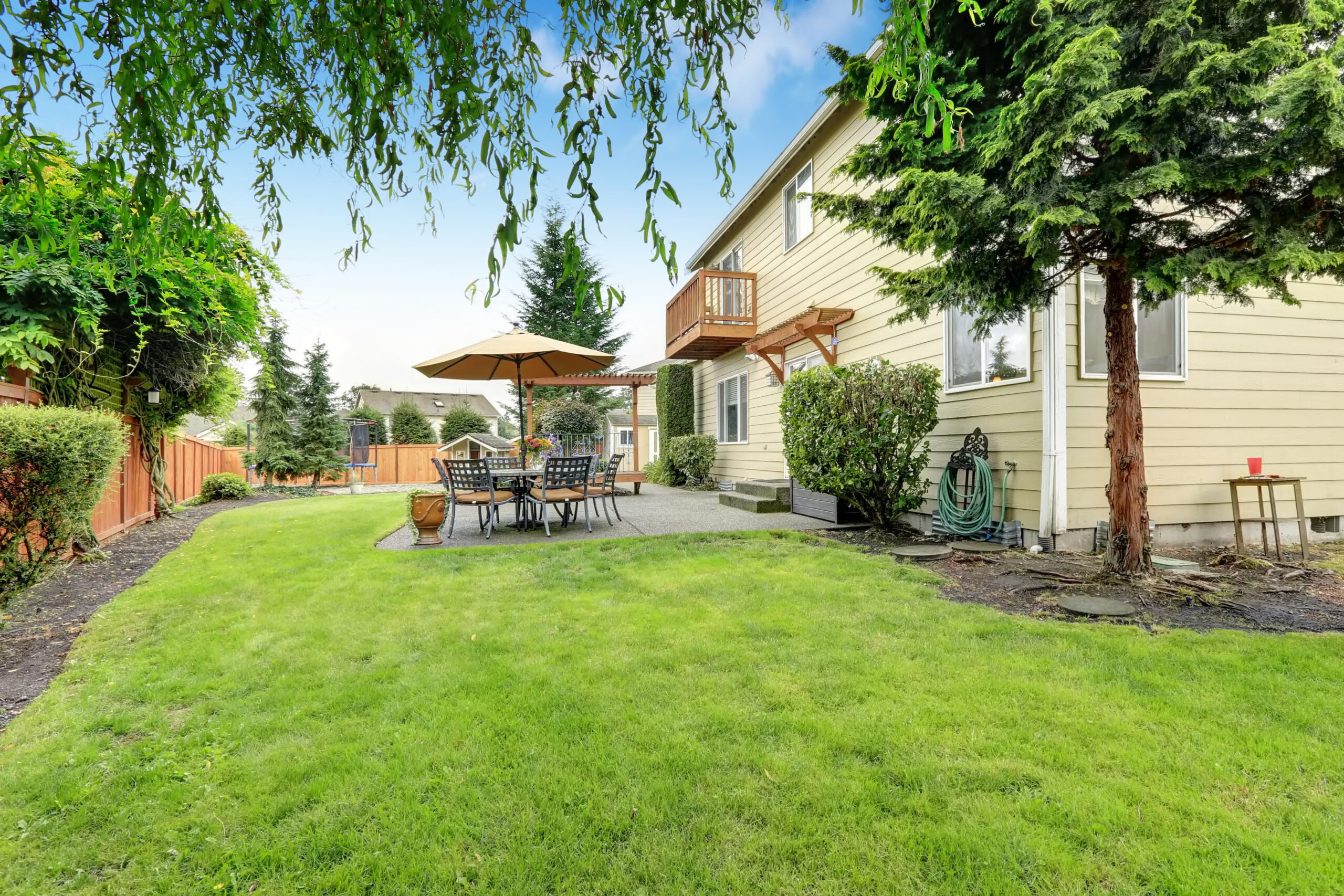 Backyard patio with dining set under umbrella beside well-maintained lawn, surrounded by trees, fencing, and garden accents.