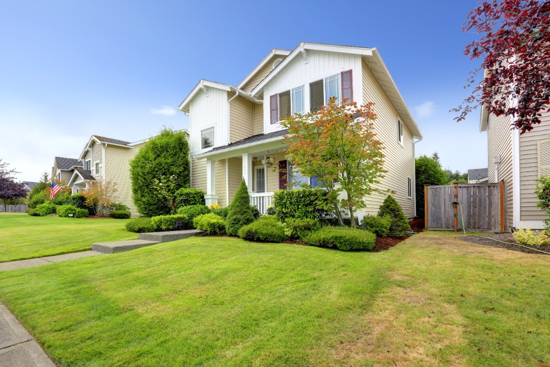 Modern beige house with red shutters and manicured front lawn, surrounded by lush shrubs, trees, and a clear blue sky above.